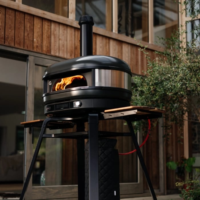 Black outdoor pizza oven with flames inside, placed on a stand outdoors.