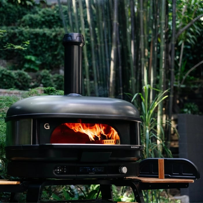 Black outdoor pizza oven with fire inside, set against a natural background of greenery.