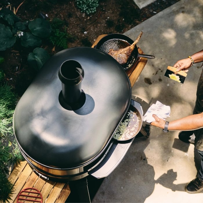 Person cooking outdoors using a black pizza oven with a lid.