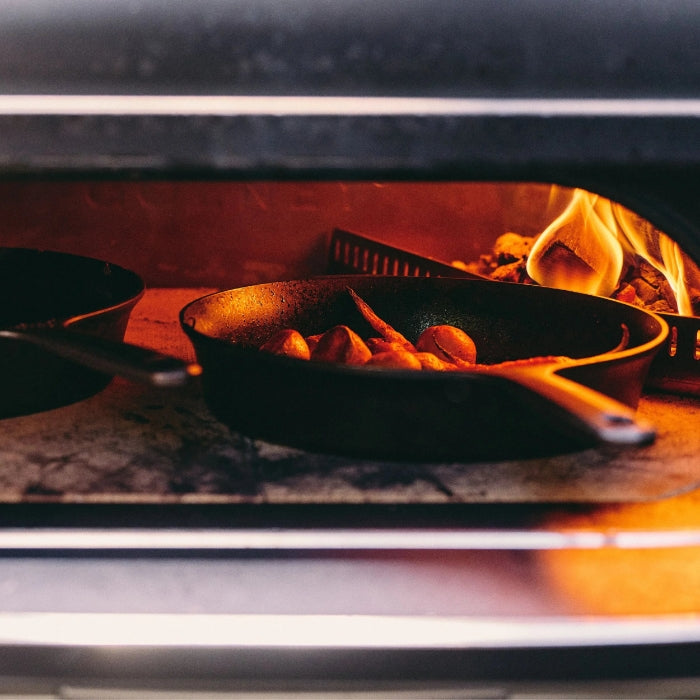 Frying pan with food being cooked over an open flame inside a wood-fired oven.