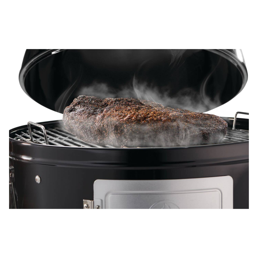A large cooked steak sits on the grill grate inside a black round smoker or grill, with visible steam rising from the meat against a white background.